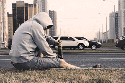 Eine Person sitzt alleine am Straßenrand mit einer Flasche Bier